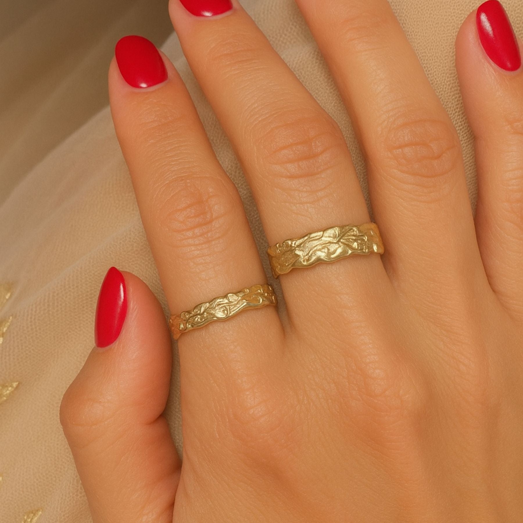 Close-up of a hand wearing two gold rings with red nail polish on a beige fabric background